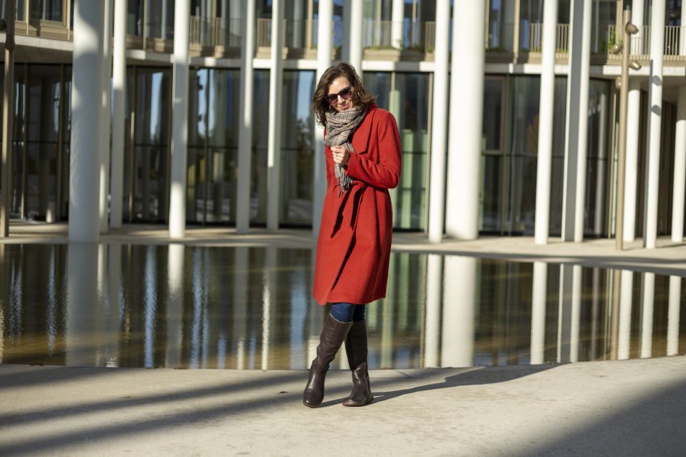 Eine Frau im roten Mantel steht vor einem modernen Glasgebäude mit hohen weißen Säulen. Sie trägt eine Sonnenbrille, einen Schal, Jeans und stylische kniehohe Stiefel in der Farbe QATCH Winter Brown von ZAQQ. Sie posiert auf einer sonnenbeschienenen Terrasse in der Nähe eines reflektierenden Wasserspiels.