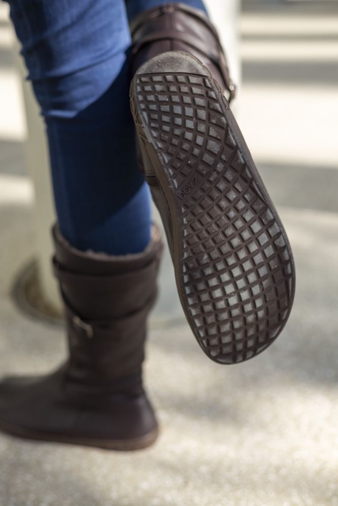 Eine Person, die die ATTRAQT Winter Brown Stiefel von ZAQQ und Blue Jeans trägt, hebt den linken Fuß und gibt den Blick auf die strukturierte Sohle frei, die hervorragenden Tragekomfort verspricht. Der Hintergrund ist eine helle Fläche.