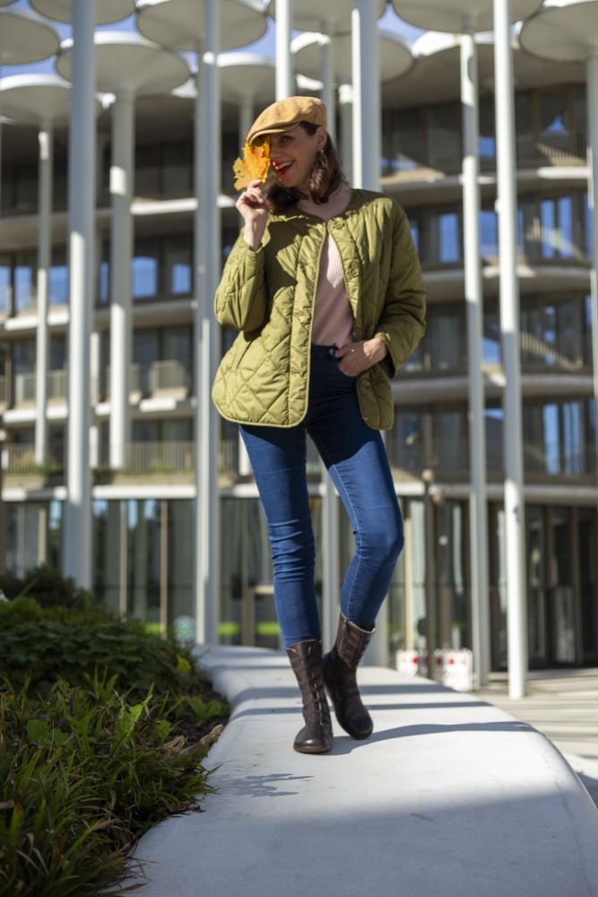 Eine Person in grüner Steppjacke und Blue Jeans steht auf einer gewölbten Steinfläche und trägt den flexiblen ATTRAQT Stretch Nappa Brown von ZAQQ. Sie hält ein Blatt mit Gebäuden im Hintergrund und trägt eine beige Mütze. Sie lächelt unter blauem Himmel in die Kamera.