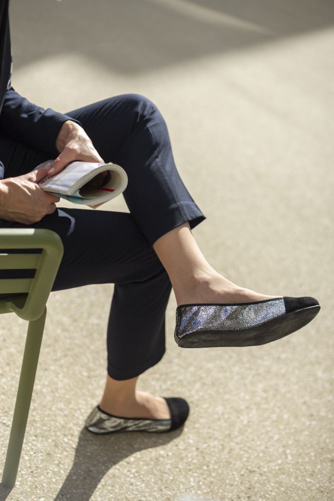 Eine Person sitzt auf einem grünen Stuhl, trägt marineblaue Hosen und schwarze TWIST Derby-Schuhe von ZAQQ und hält eine aufgeschlagene Zeitschrift in der Hand. Die Szene scheint im Freien zu sein, wobei das Sonnenlicht Schatten auf den Boden wirft.