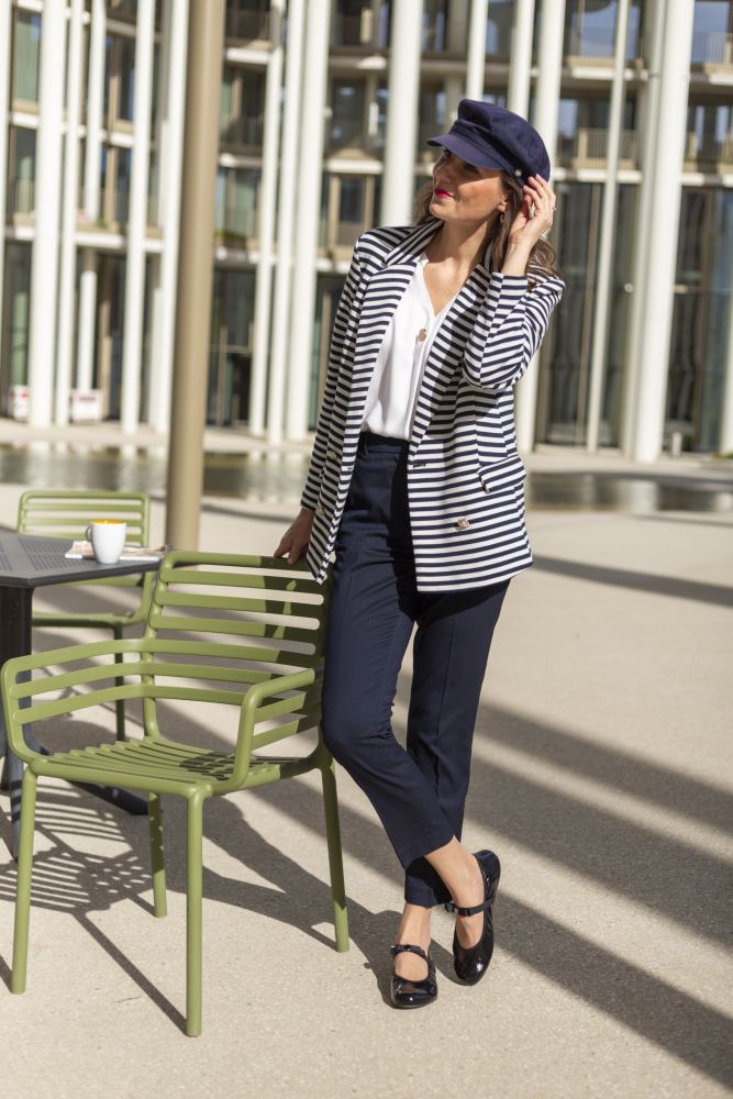 Eine Frau mit marineblauer Mütze und gestreiftem Blazer steht neben einem grünen Tisch und Stühlen im Freien. Sie trägt eine weiße Bluse, marineblaue Hosen und TWIST CASEMIRO Schuhe von ZAQQ mit flexiblen Sohlen. Sonnenlicht wirft starke Schatten auf den Boden. Auf dem Tisch neben ihr steht eine Tasse.