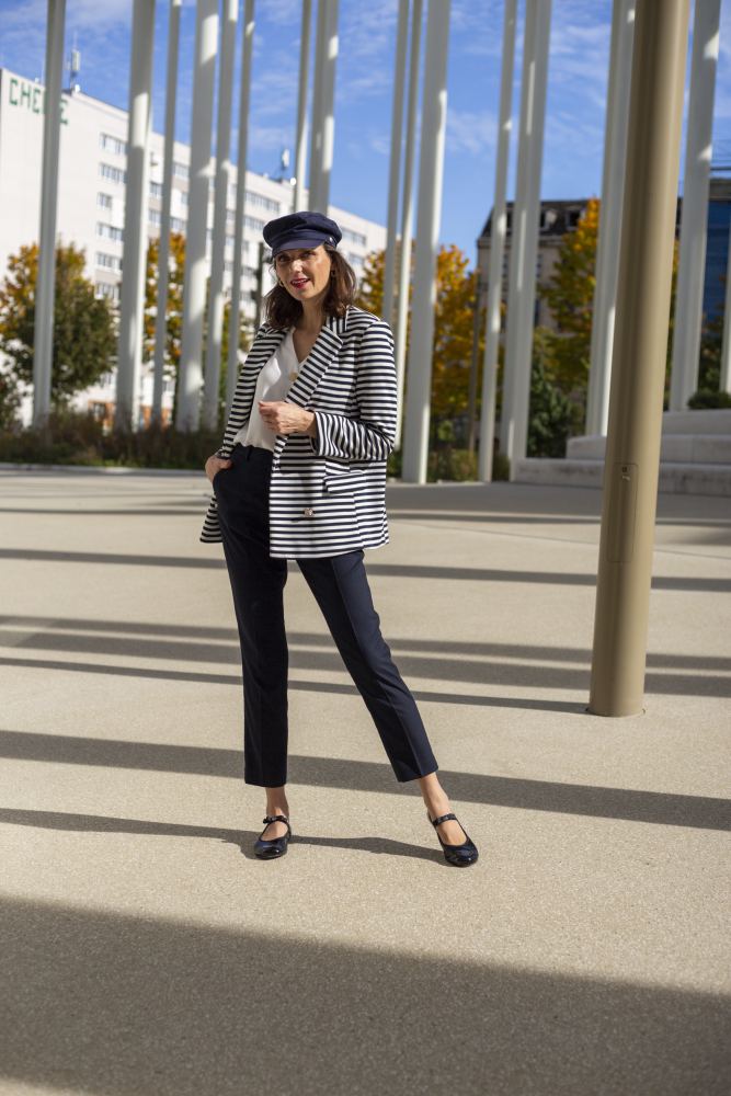 Eine Frau posiert selbstbewusst im Freien vor hohen Säulen. Sie trägt einen gestreiften Blazer, eine schwarze Hose und eine marineblaue Mütze. Ihre TWIST CASEMIRO Schuhe von ZAQQ mit flexibler Sohle und bequemem Design lassen sie entspannt aussehen. Der klare blaue Himmel und die Herbstbäume vervollständigen die Szene mit Schatten, die auf den Boden geworfen werden.