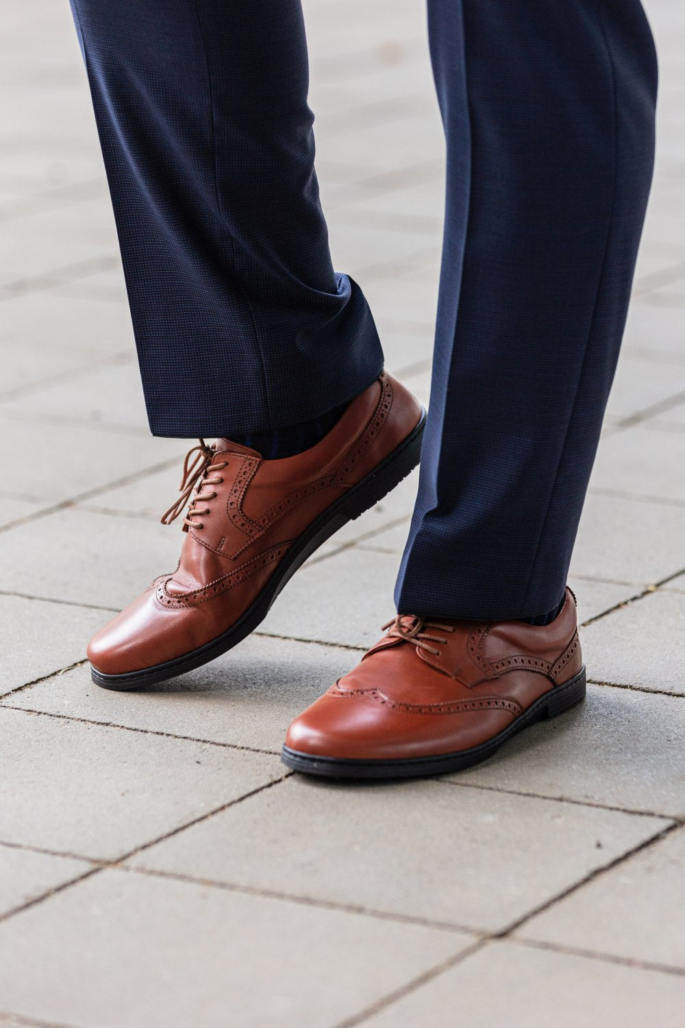 Eine Person in dunkelblauen Hosen und braunen Brogue-Schuhen aus Leder der Linie BRIQ Brogue Cognac von ZAQQ steht auf einem gefliesten Bürgersteig. Diese stilvollen Schuhe sind mit dekorativen Perforationen und Schnürsenkeln versehen und ergänzen die formelle Kleidung auf wunderbare Weise.