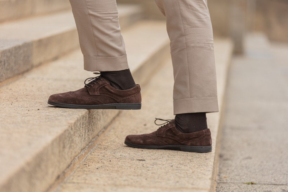 Eine Person in beiger Hose und den Schuhen „BRIQ Brogue Velours Brown“ von ZAQQ betritt eine Steintreppe.