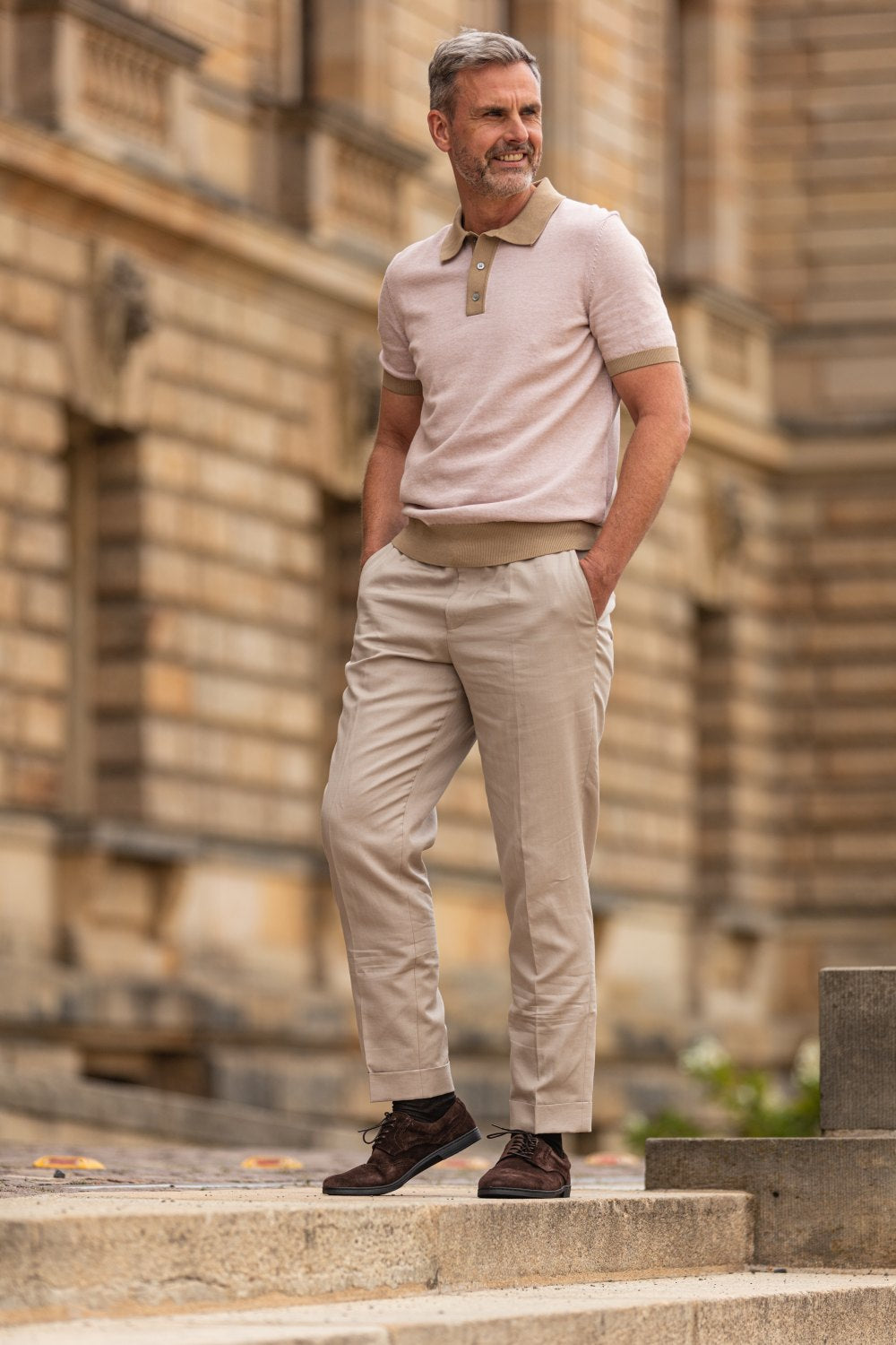 Ein Mann mit grauem Haar und Bart steht auf Steinstufen vor einem historischen Gebäude. Er trägt ein beiges Poloshirt und eine beige Hose, ergänzt durch die BRIQ Brogue Velours Brown von ZAQQ. Er sieht entspannt und zufrieden aus und strahlt klassische Eleganz aus.