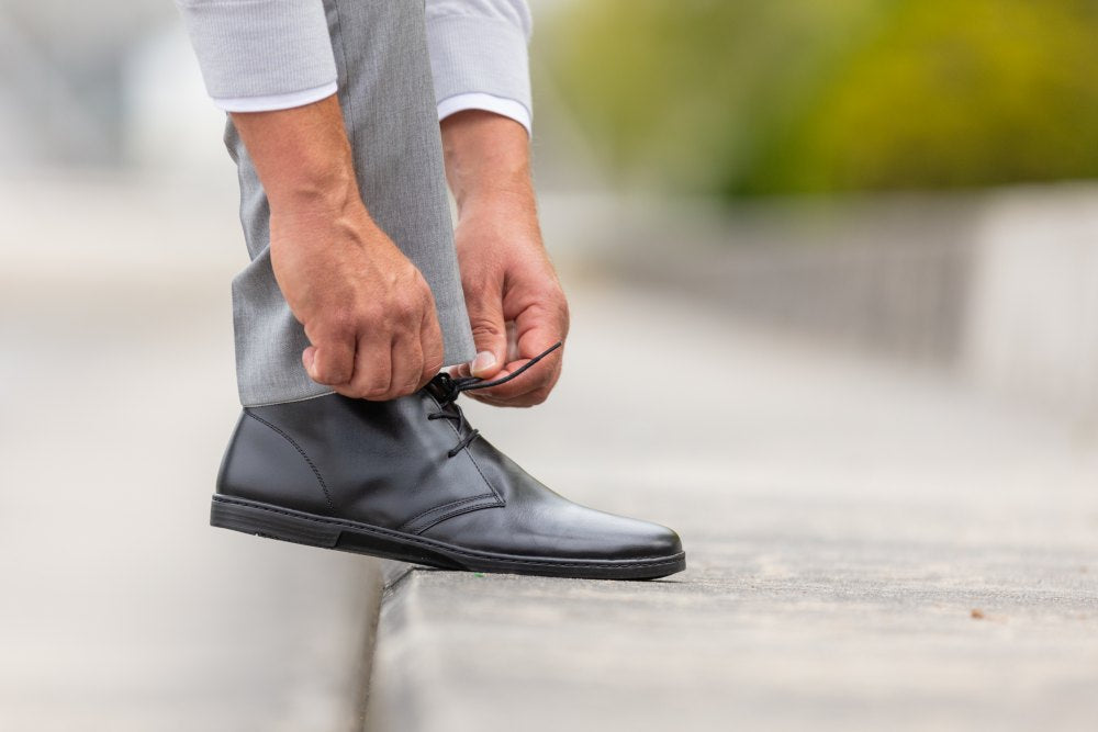 Eine Person in grauen Hosen und weißem Hemd bückt sich, um die Schnürsenkel eines schwarzen DUQE-Stiefels von ZAQQ zu binden, während sie auf einem Betonvorsprung steht, vor einem unscharfen Hintergrund mit Grünpflanzen.