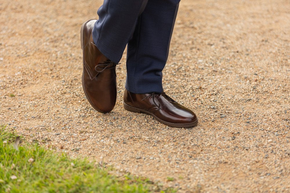 Eine Person, die die Schuhe DUQE Vegan Antique Brown von ZAQQ und eine dunkelblaue Hose trägt, geht auf einem Kiesweg in der Nähe eines Stücks grünem Gras.