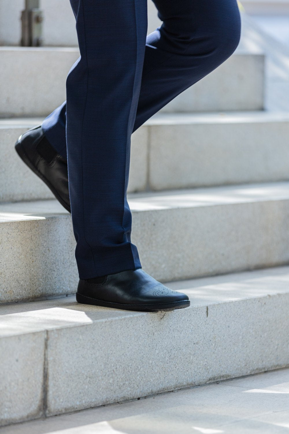 Eine Person in dunkelblauen Hosen und eleganten schwarzen ZAQQ EQUITY Brogue-Schuhen geht anmutig im hellen Licht Steintreppen hinunter.