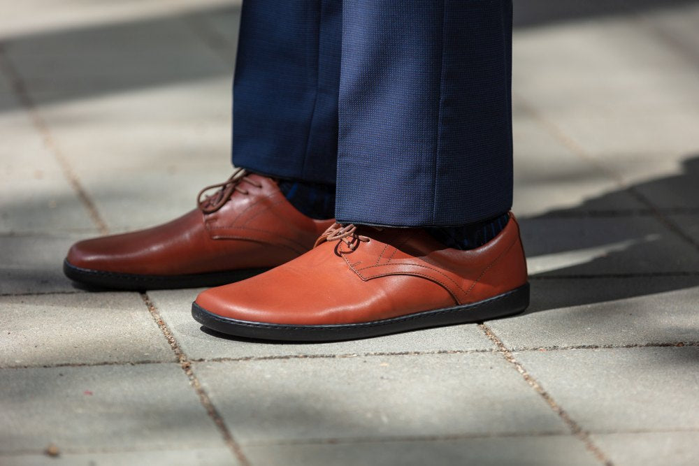 Eine Person trägt ZAQQs PEAQ Cognac-braune Lederschuhe mit Barfußsohle und dunkelblaue Hosen und steht auf einem gefliesten Gehweg. Die Glattlederschuhe sind ordentlich geschnürt und das Sonnenlicht wirft weiche Schatten auf die Fliesen.