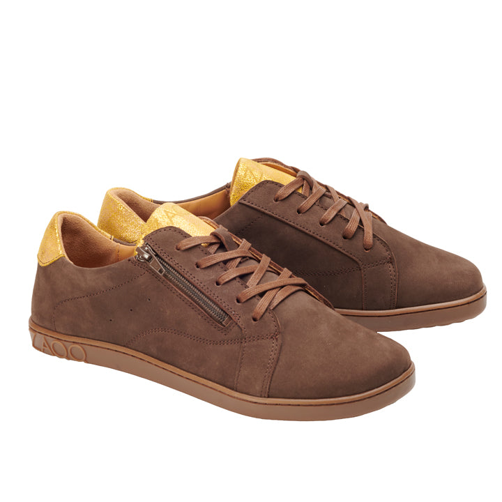 Wir präsentieren den QOMFY Wide Brown von ZAQQ: ein Paar braune Wildleder-Sneaker mit goldenen Akzenten an Ferse und Zunge, gefertigt für optimalen Komfort. Diese Schuhe verfügen über passende braune Schnürsenkel, einen dekorativen seitlichen Reißverschluss und eine stützende Sohle, die das Gesamtfarbschema perfekt ergänzt.