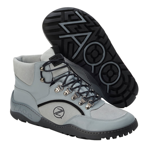 Eine Nahaufnahme der TREQ Grey Waterproof Wanderstiefel von ZAQQ, die ihr stilvolles graues Erscheinungsbild mit schwarzen Sohlen zeigt. Die Stiefel sind mit Metallösen und schwarzen Schnürsenkeln ausgestattet und tragen das ZAQQ-Logo sowohl an der Seite als auch an der Sohle. Ein Stiefel steht aufrecht, während der andere geneigt ist, um sein Sohlenmuster zu zeigen.