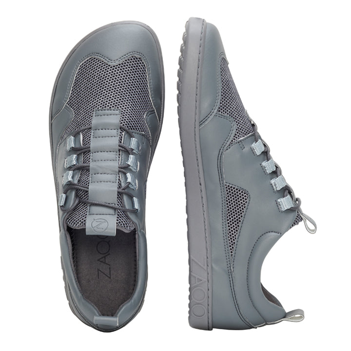 Wir präsentieren den QARLEE Grey von ZAQQ: ein minimalistisches Paar Sneaker mit schlichtem Design. Der linke Schuh ist von oben zu sehen, während der rechte Schuh von der Seite zu sehen ist. Beide Schuhe sind mit einem Obermaterial aus Mesh und einer flexiblen Sohle ausgestattet und bieten so einen Komfort, der mit dem eines Barfußschuhs vergleichbar ist.