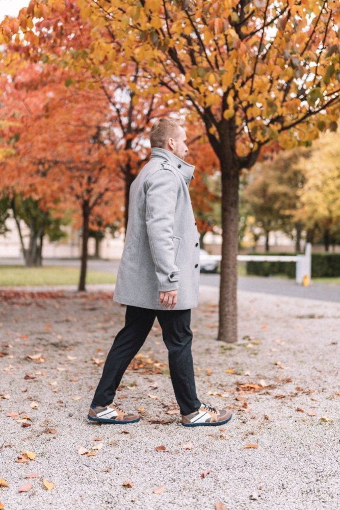 Eine Person in einem grauen Mantel geht einen Kiesweg entlang, der von Bäumen gesäumt ist, die mit leuchtenden Herbstblättern in Orange- und Rottönen geschmückt sind. Die parkähnliche Umgebung ist ruhig, überall liegen gefallene Blätter verstreut, während die Person einen Spaziergang in BREAQ Sierra-Sneakern von ZAQQ mit der flexiblen ZAQQ-Sohle genießt.