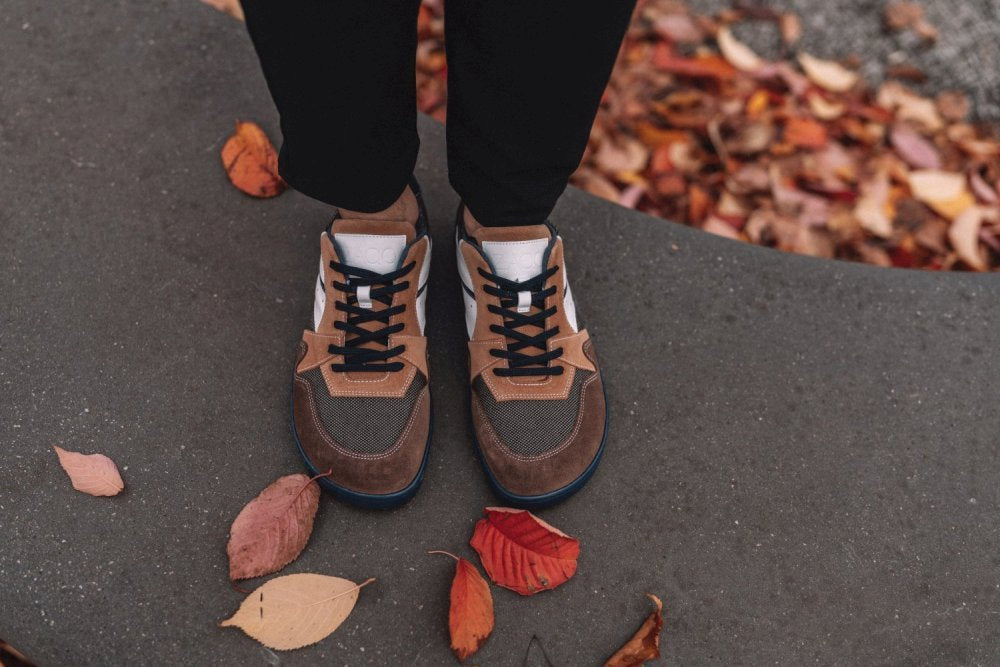 Eine Person, die ZAQQs BREAQ Sierra Sneaker mit schwarzen Schnürsenkeln trägt, steht auf einer grauen Betonfläche. Um sie herum sind gefallene Herbstblätter in Rot-, Orange- und Brauntönen verstreut, die den Stil und den Komfort dieser Barfußschuhe betonen.