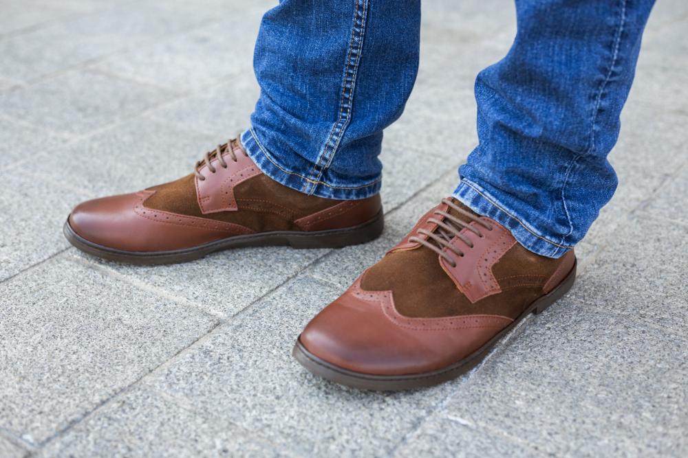 Nahaufnahme einer Person, die BRIQ Brogue Antique Cognac-Schuhe von ZAQQ trägt, in Braun- und Olivgrüntönen gehalten, kombiniert mit Blue Jeans. Diese Kalbsleder-Oxford-Schuhe zeichnen sich durch eine Mischung aus Leder- und Wildledertexturen aus, ideal als Businessschuh, während die Person auf einem grau gefliesten Bürgersteig steht.