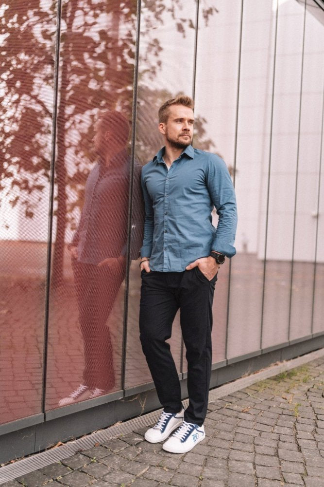 Ein Mann in einem CEDRIQ-blauen Hemd von ZAQQ und einer schwarzen Hose lehnt an einem modernen Glasgebäude. Er steht auf einem Kopfsteinpflasterweg und trägt elegante Barfußschuhe mit blauen Akzenten. Das Glas spiegelt die umliegenden Bäume und verleiht der städtischen Umgebung ein natürliches Element.