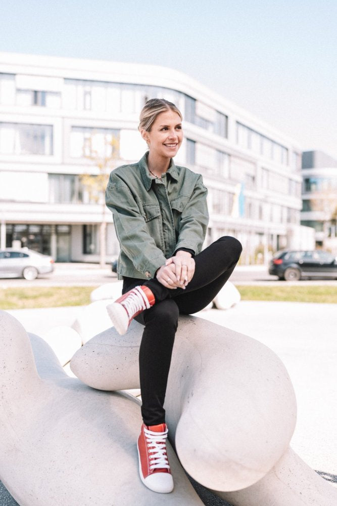 Eine Person mit kurzen blonden Haaren sitzt auf einer großen, abstrakten weißen Skulptur im Freien. Sie trägt eine grüne Jacke, eine schwarze Hose und rote ZAQQ CHUQQS-Sneaker. Im Hintergrund sind ein modernes Gebäude und geparkte Autos zu sehen.