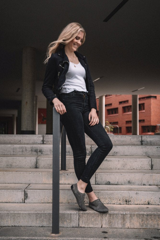 Eine Person mit langen, welligen Haaren, einer schwarzen Jacke, einem weißen Hemd und Jeans lehnt an einem Geländer einer Betontreppe. Im Hintergrund sind rote Backsteingebäude zu sehen, die stilvoll den IQON Velours Grey von ZAQQ tragen.