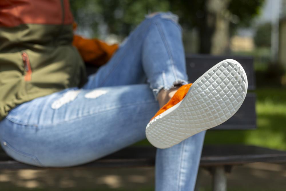 Eine Person sitzt mit übereinandergeschlagenen Beinen auf einer Bank. Sie trägt zerrissene blaue Jeans, LIQE Orange-Schuhe von ZAQQ mit weißer Sohle, die ein Barfußgefühl vermittelt, und eine grüne Jacke. Der Fokus liegt auf der Schuhsohle, umgeben von üppigem Grün im Hintergrund.