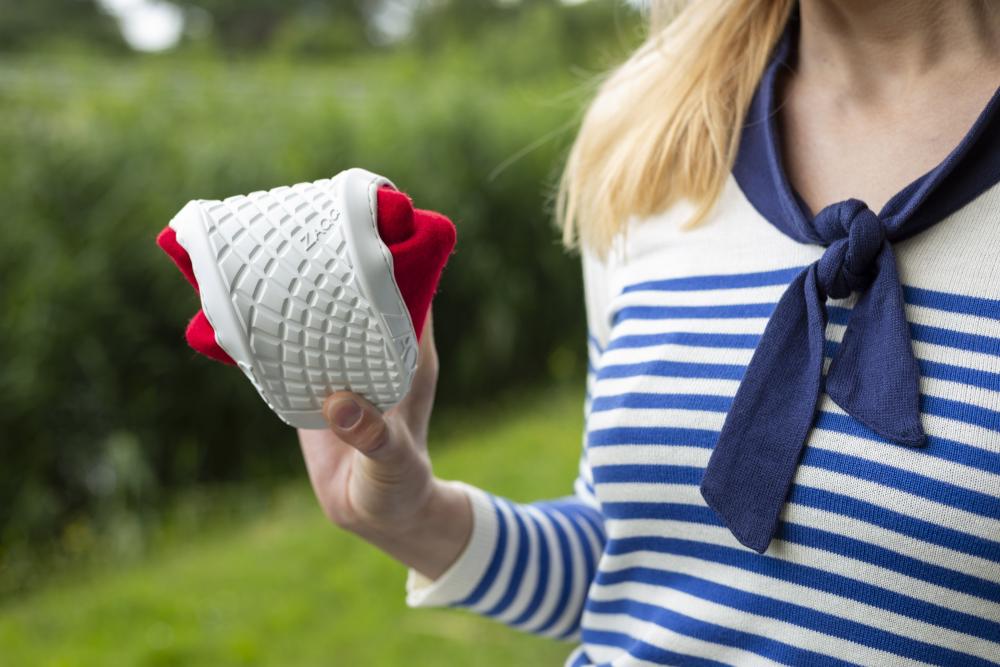 In einer Außenkulisse mit Grün im Hintergrund hält eine Person in einem blau-weiß gestreiften Oberteil einen aufgerollten LIQE Race-Sneaker von ZAQQ mit einer Sohle mit Waffelmuster ähnlich der Barfußsohle und einer roten Innenausstattung.