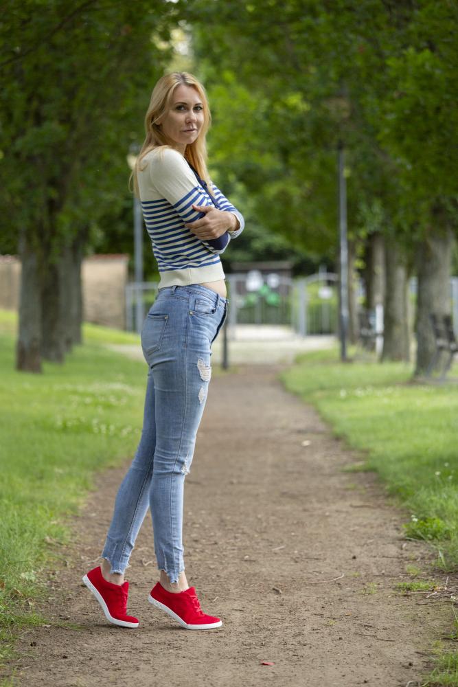 Eine Frau steht mit verschränkten Armen auf einem Weg in einem von grünen Bäumen umgebenen Park. Sie trägt einen gestreiften Pullover, zerrissene Jeans und leuchtende LIQE Race-Sneaker mit einem Hauch Rot von ZAQQ.