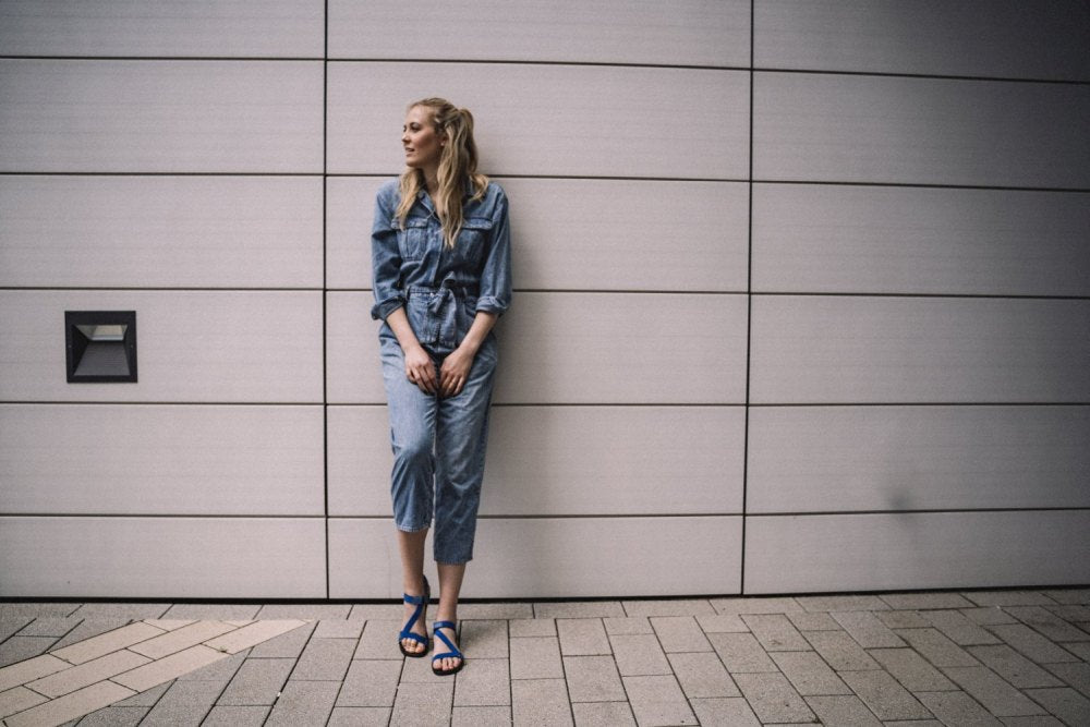 Eine Frau in einem Jeansoverall und den MIQA Blue Sandalen von ZAQQ mit Kautschuksohle steht vor einer hellgrau gefliesten Wand. Sie blickt zur Seite, die Hände vor sich gefaltet, steht auf einem hellen Ziegelpflaster.