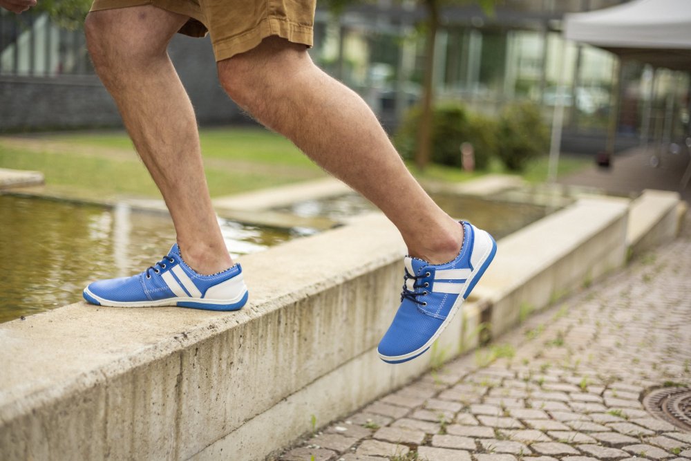 Eine Person, die die blauen QANBERRA-Sneakers und braunen Shorts von ZAQQ trägt, schlendert am Rand einer niedrigen Mauer in der Nähe eines reflektierenden Pools entlang. Die Szene spielt sich vor einem Hintergrund aus Gras, Pflaster und verschwommenen Gebäuden ab.