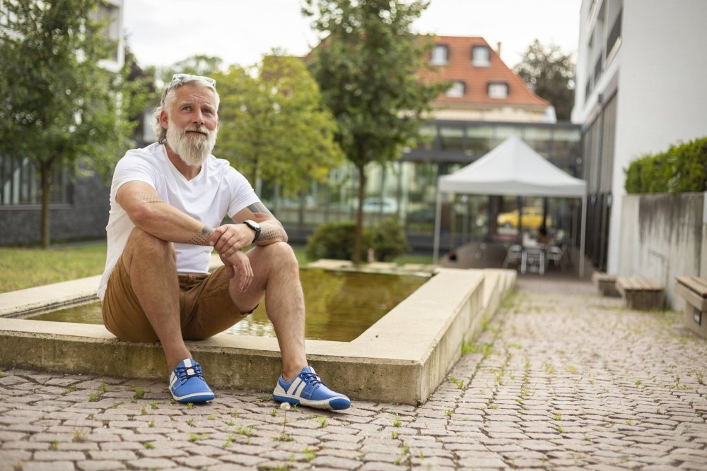 Ein Mann mit Bart und weißem Haar sitzt am Rand eines Brunnens. Er trägt ein weißes T-Shirt, braune Shorts und ein Paar Barfußschuhe QANBERRA Blue von ZAQQ. Er steht draußen auf einer gepflasterten Fläche mit Bäumen und Gebäuden im Hintergrund.