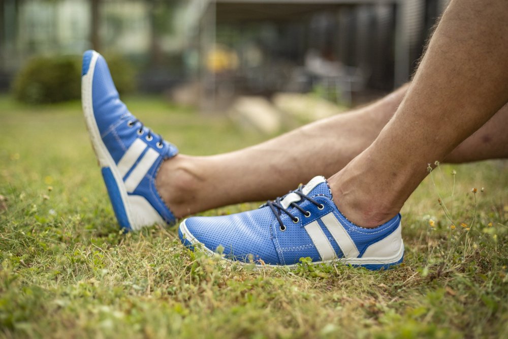 Eine Nahaufnahme zeigt eine Person, die auf Gras sitzt und die mit weißen Streifen verzierten QANBERRA Blue-Sneakers von ZAQQ präsentiert, die fachmännisch aus hochwertigem Nappaleder gefertigt wurden. Das Bild ist nur von den Beinen abwärts sichtbar und steht vor einer sanft verschwommenen Gartenkulisse, die lässige Eleganz ausstrahlt.