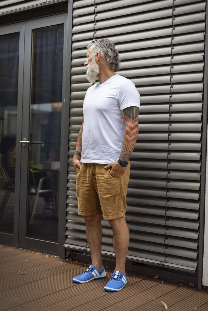 Ein Mann mit weißem Bart und Tattoos steht vor einem Gebäude mit grauen Lattenwänden. Er trägt ein QANBERRA Blue T-Shirt von ZAQQ, dazu braune Shorts und blaue Barfußschuhe aus hochwertigem Nappaleder, blickt zur Seite und hat die Hände in den Taschen.