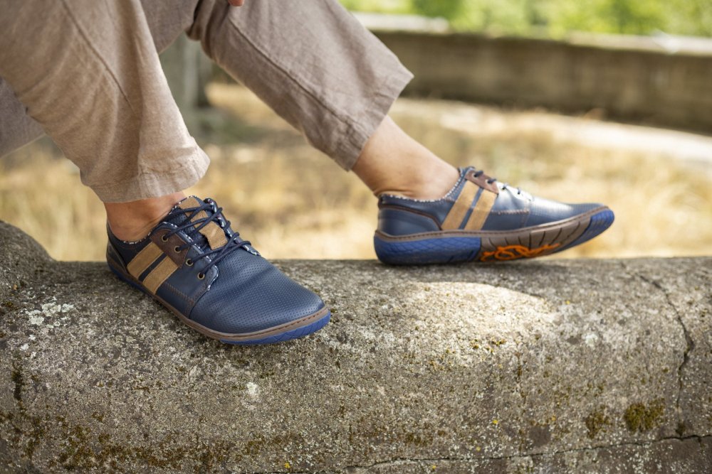 Eine Person trägt QANBERRA Navy-Schuhe von ZAQQ mit minimalistischem Sohle-Design und sitzt in hellbraunen Hosen auf einem Felsvorsprung. Die Szene ist vor einem Hintergrund mit verschwommenem Grün und einer Wand angesiedelt.