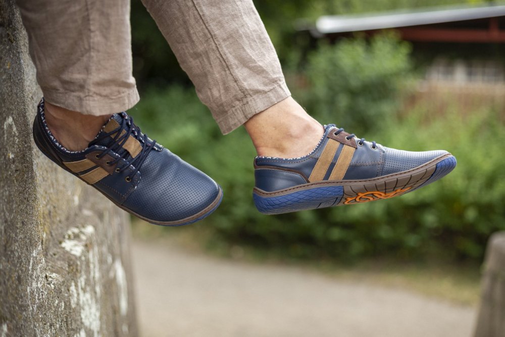 Eine Nahaufnahme der Füße einer Person, die die eleganten QANBERRA Navy-Schuhe von ZAQQ trägt, hergestellt aus hochwertigem Nappaleder in einem stilvollen blau-braunen Barfußschuh-Design mit einer minimalistischen Sohle. Die Person sitzt auf einem Felsvorsprung, mit verschwommenem Grün und einem Gebäude im Hintergrund.