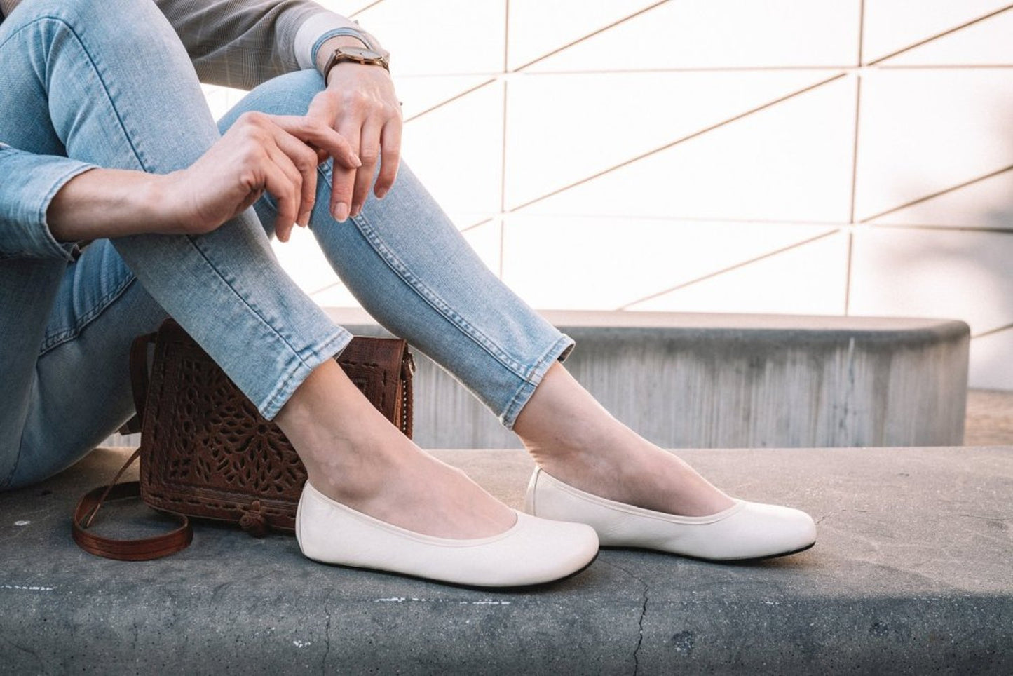 Eine Person in hellblauer Jeans und QARMA Beige Ballerinas von ZAQQ sitzt auf einer Betonbank. Ihre Hände ruhen auf einer braunen Lederhandtasche mit dekorativem Cut-Out-Design. Im Hintergrund ist eine moderne, geometrische weiße Wand zu sehen.