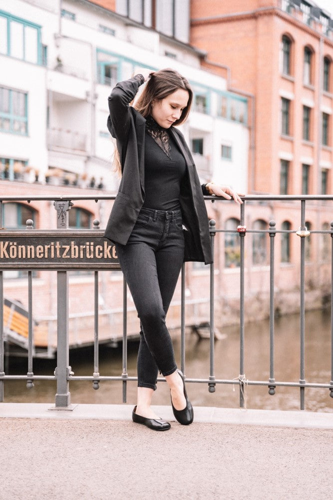 Eine Frau in QARMA Black von ZAQQ steht auf der Könneritzbrücke, einen Fuß elegant über den anderen geschlagen wie eine Ballerina. Die Gebäude im Hintergrund verleihen ihrer anmutigen Haltung einen urbanen Touch.