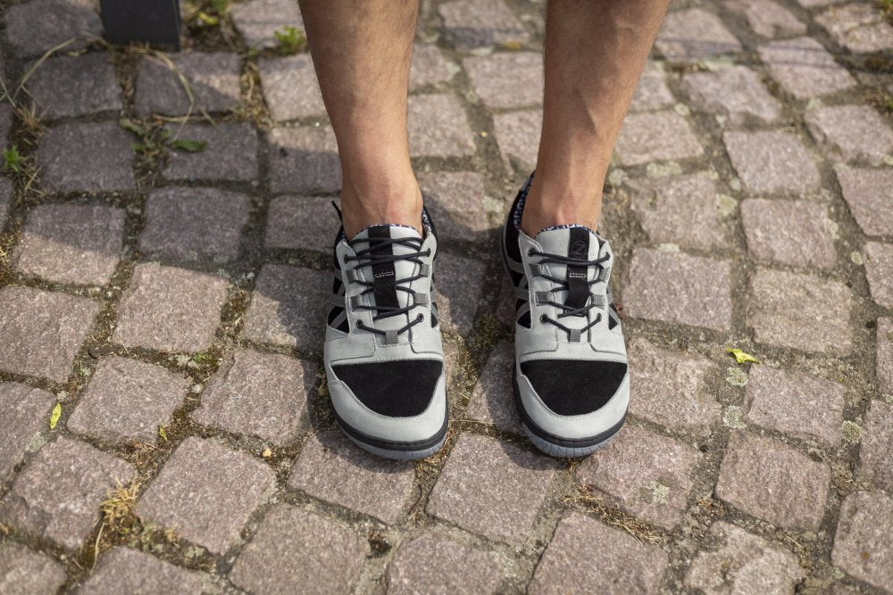 Eine Person steht in den stylischen QENDRIQ Black Grey Sneakers auf einem Kopfsteinpflasterweg. Nur Beine und Schuhe sind zu sehen, was das minimalistische Design dieses Sneakers von ZAQQ mit MINIMAL-Sohle unterstreicht.