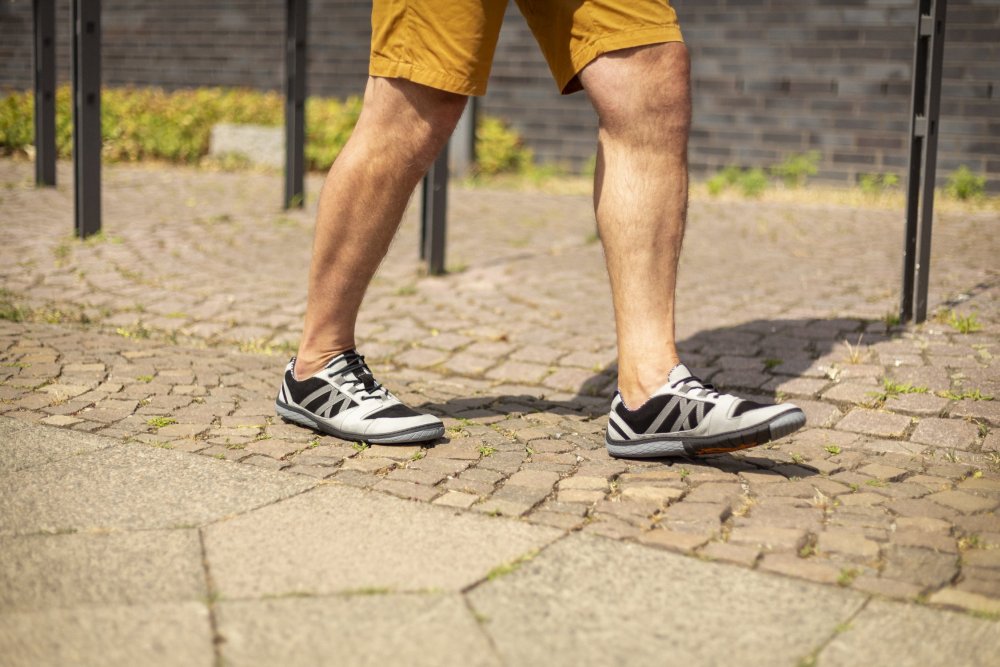 Eine Person in orangefarbenen Shorts und eleganten schwarz-grauen ZAQQ QENDRIQ-Sneakern schlendert einen Kopfsteinpflasterweg entlang, der von üppigem Grün und einer rustikalen Backsteinmauer gesäumt ist.