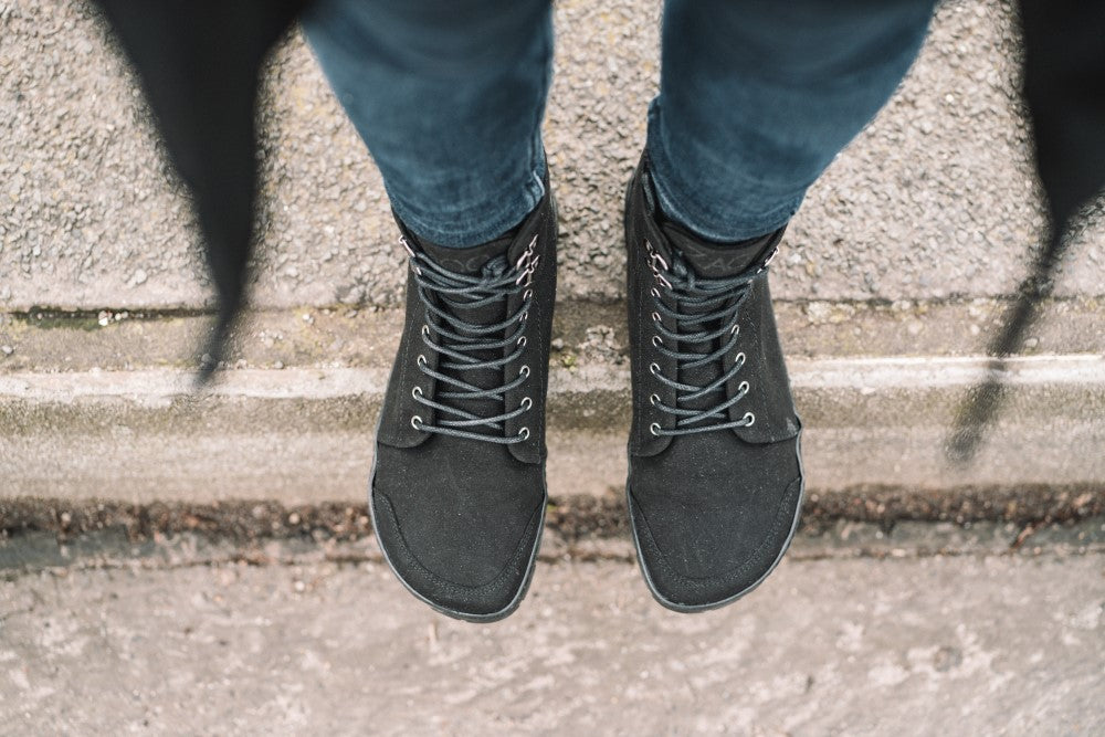 Eine Person in Blue Jeans und den schwarzen QICE High-Top-Sneakers von ZAQQ steht auf einem strukturierten Gehweg. Die Schuhe haben schwarze Schnürsenkel und das Foto zeigt eine Draufsicht der Füße vor einem rauen Betonhintergrund.