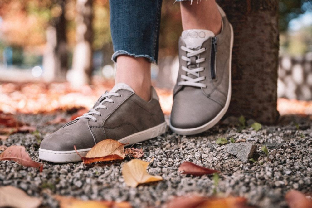 Eine Person in den QOMFY Wide Grey Sneakers von ZAQQ mit weißen Schnürsenkeln steht auf Kies, umgeben von verstreutem Herbstlaub. Ein Fuß ruht auf einem Baumstamm. Der Komfort Barfußschuh-Stil ergänzt die Blue Jeans, während der Hintergrund verschwommenes Herbstlaub zeigt.