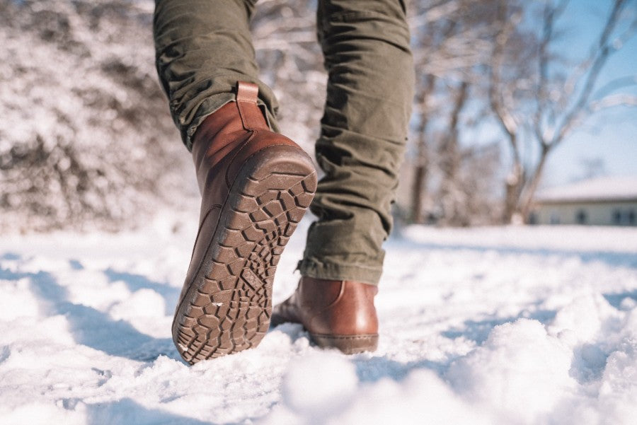 Nahaufnahme einer Person, die ZAQQ QUINTIC Winter Vegan Nut-Stiefel und grüne Hosen trägt und auf einem schneebedeckten Pfad läuft. Die Sonne scheint hell und wirft Schatten auf den knirschenden Schnee, während im Hintergrund kahle Bäume stehen.