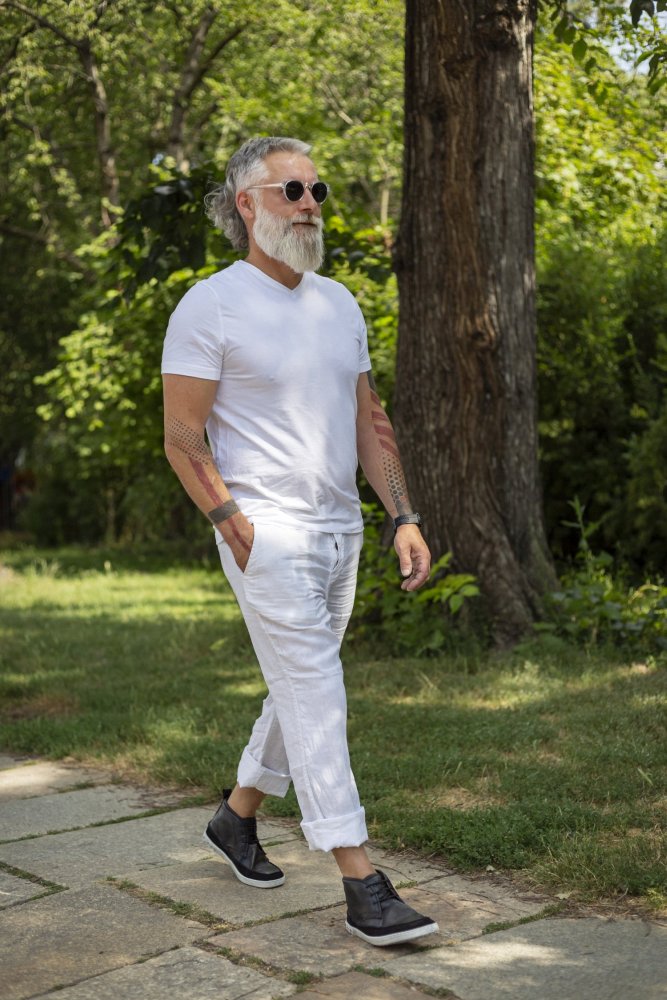 Ein Mann mit Sonnenbrille und weißem Bart schlendert stilvoll durch den Park. Er trägt Barfußsneakers, ein weißes Hemd und eine weiße Hose, ergänzt durch die Schuhe QUITMAN Black von ZAQQ. Grüne Bäume und Gras umgeben ihn, während im Hintergrund stolz ein großer Baumstamm thront.