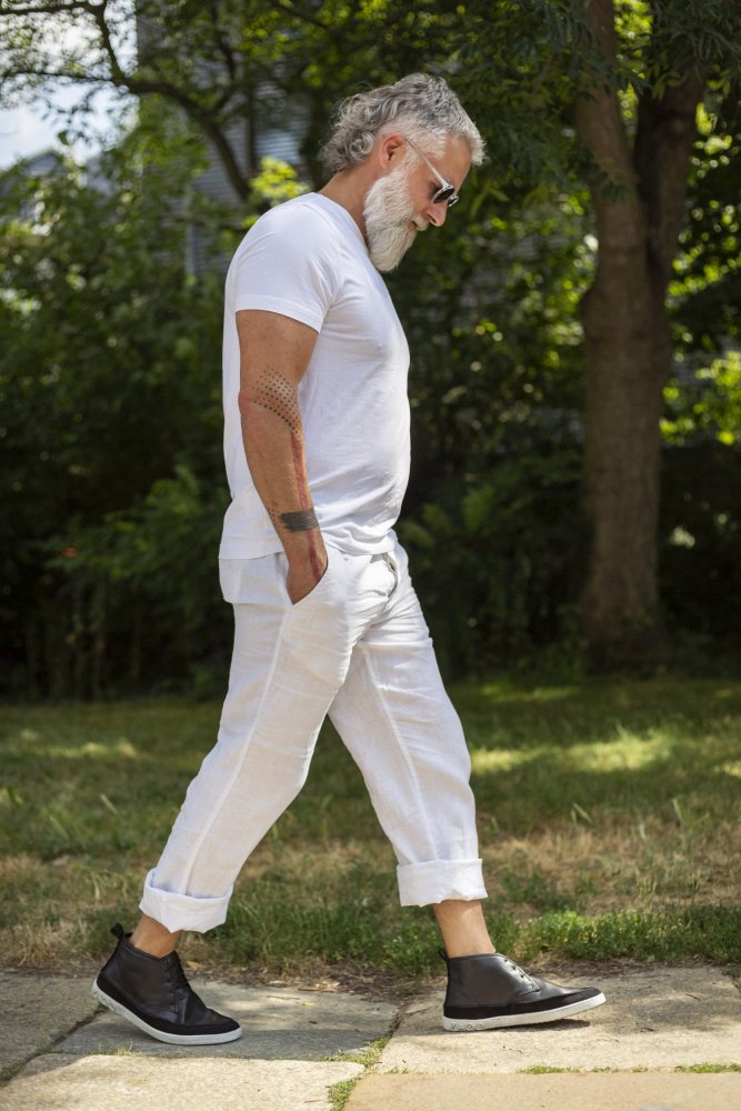 Ein bärtiger Mann in weißem T-Shirt und hochgekrempelter Hose spaziert an einem sonnigen Tag draußen herum. Er trägt eine stylische Sonnenbrille und hat die Hände in den Taschen. Seine QUITMAN Black Sneakers von ZAQQ aus glattem Nappaleder verleihen dem Hintergrund aus üppigen Bäumen und Gras einen Hauch von Flair.