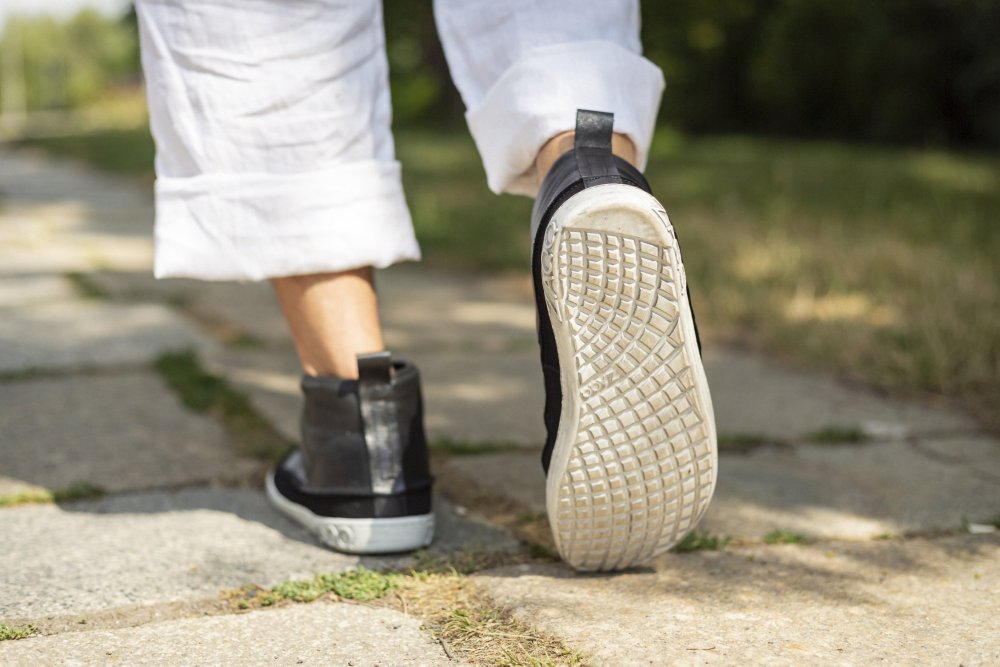 Eine Person in stylischen QUITMAN Black Barfuß-Sneakers von ZAQQ schlendert einen Steinweg entlang. Ihre weiße Hose ist über die Knöchel hochgekrempelt. Der Weg ist von Gras und Bäumen umgeben und erinnert an eine Park- oder Gartenkulisse.