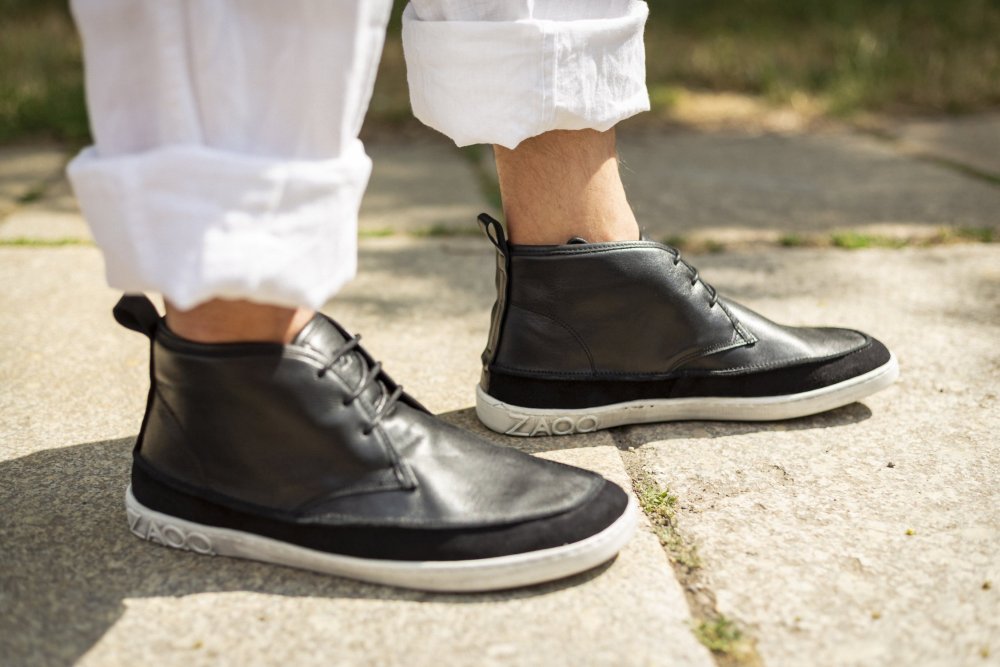 Eine Person trägt elegante schwarze QUITMAN High-Top-Sneaker von ZAQQ mit weißen Sohlen und an den Knöcheln hochgekrempelten Hosen und steht auf einem Steinweg.