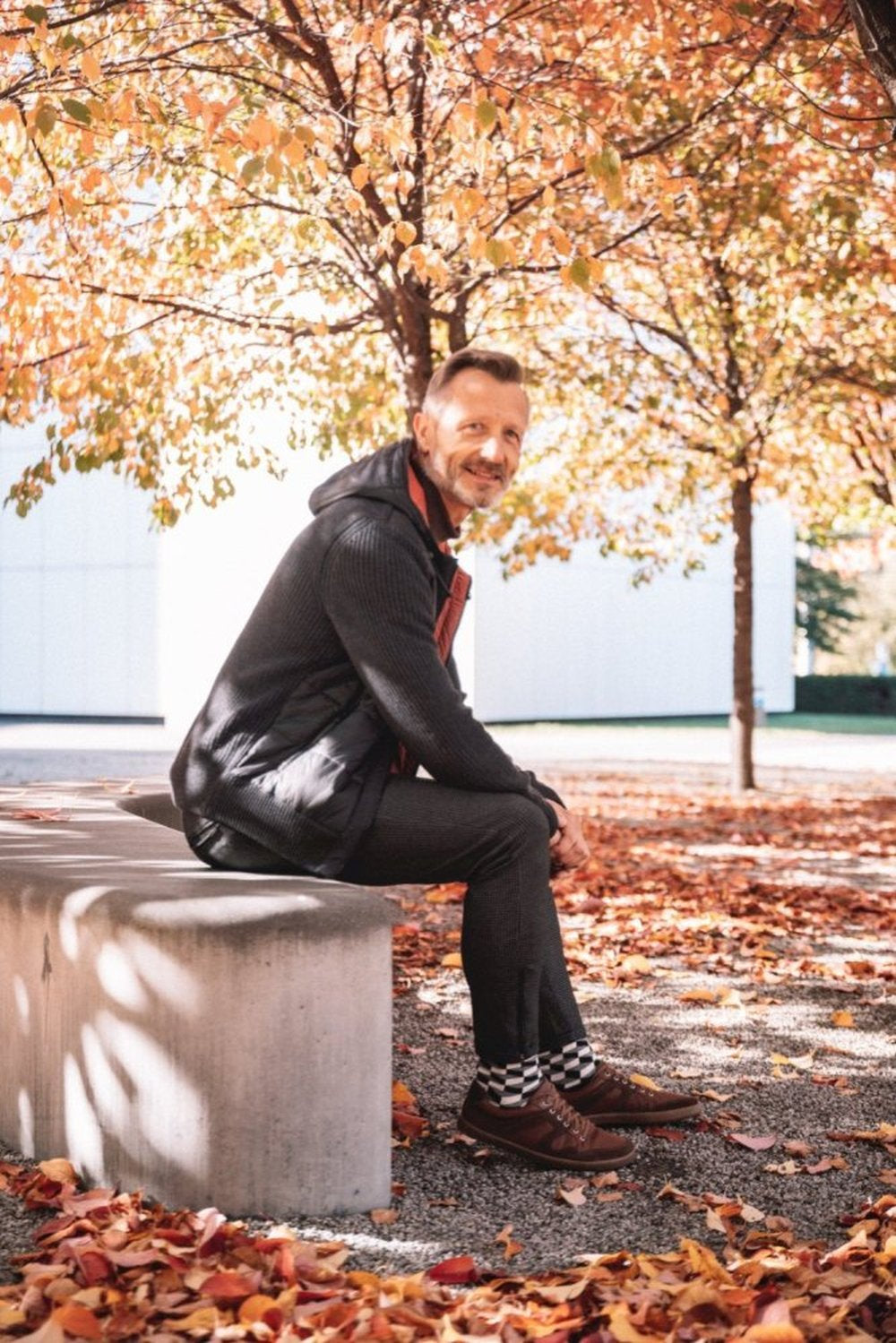 Eine Person sitzt auf einer Betonbank unter Bäumen, die voller leuchtender Herbstblätter sind. Sie trägt dunkle Kleidung und die RIQO Brown-Schuhe von ZAQQ für unvergleichlichen Komfort. Sie ist von gefallenen Blättern umgeben, in einer sonnendurchfluteten Szene mit einem weißen Gebäude im Hintergrund.