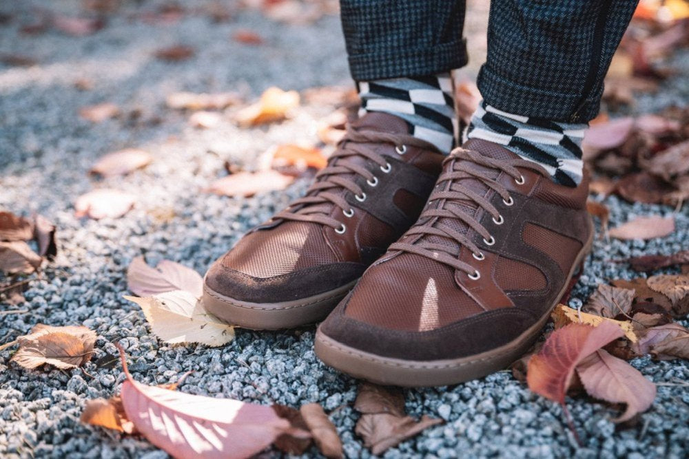 Nahaufnahme einer Person, die RIQO Brown-Sneaker von ZAQQ trägt, bekannt für ihren Tragekomfort, kombiniert mit schwarz-weiß karierten Socken. Sie steht auf einem mit trockenem Laub übersäten Kiesweg, trägt dunkle karierte Hosen und schafft so einen mühelos stylischen Look.