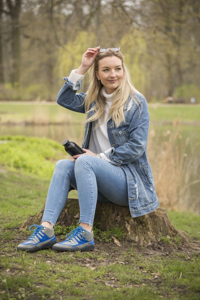 Eine Frau in Jeansjacke und Jeans sitzt auf einem Baumstumpf in einem Park und hält eine Sonnenbrille und eine schwarze Wasserflasche in der Hand. Sie blickt lässig zur Seite, hinter ihr sieht man üppiges Grün aus Gras, Bäumen und einem Teich. Ihr Outfit wird mit den SQOUT Grey Blue Waterproof-Schuhen von ZAQQ perfekt zu ihrem Barfußschuh-Stil komplementiert.