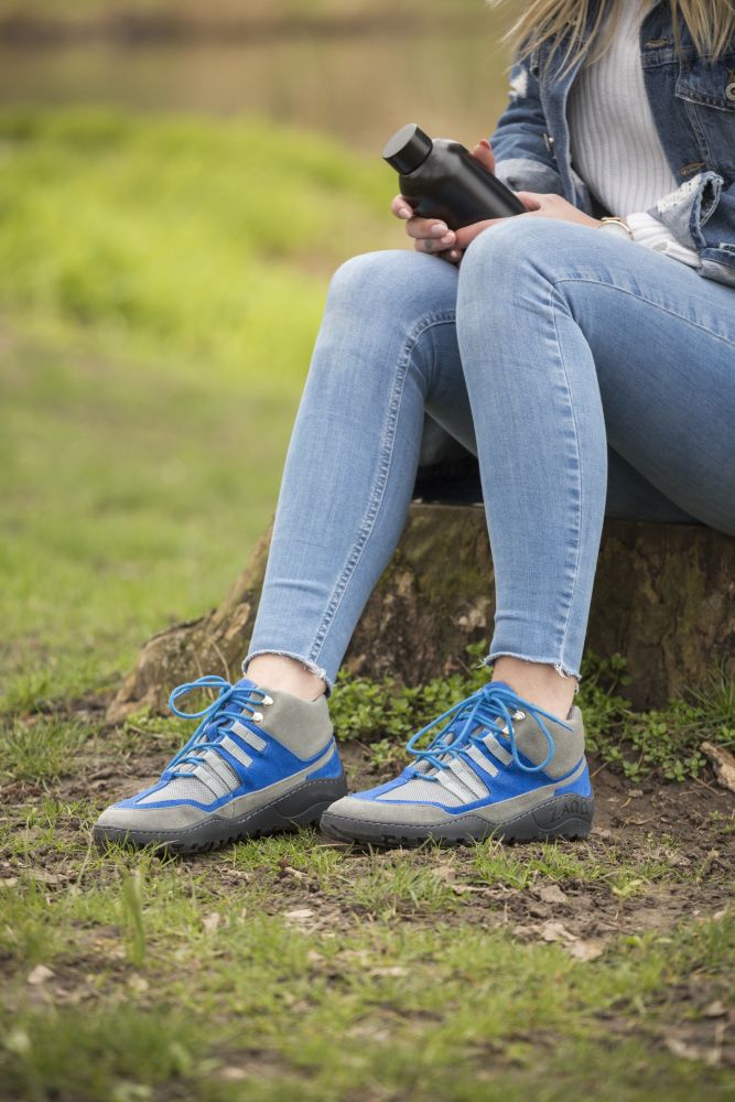 Eine Person sitzt draußen auf einer Wiese und trägt wasserfeste SQOUT Grey Blue Sneakers von ZAQQ. Sie hält eine schwarze Flasche in der Hand und sitzt auf einem Baumstumpf, umgeben von üppigem Grün und einem verschwommenen Teich im Hintergrund.