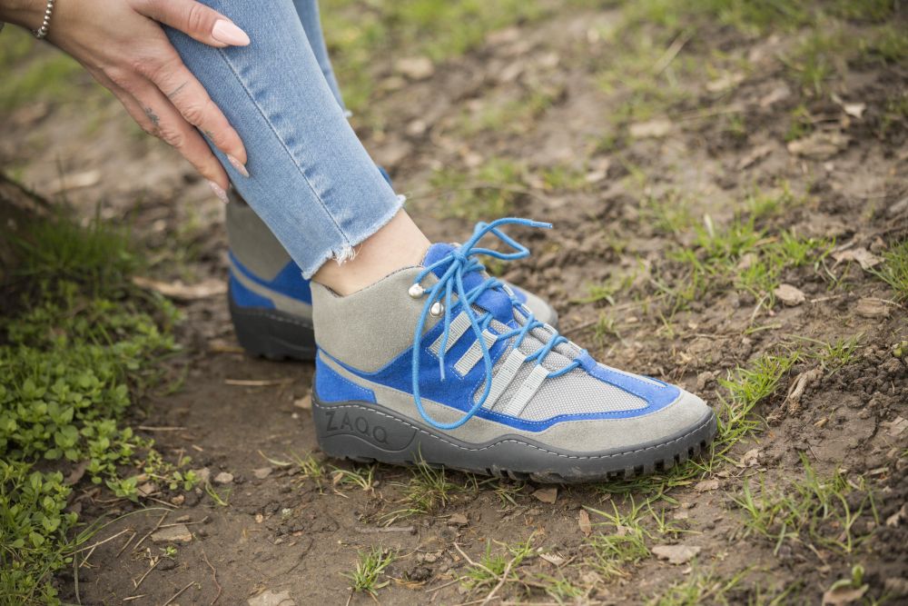 Eine Person, die SQOUT Grey Blue Waterproof Wanderschuhe von ZAQQ mit Jeans trägt, kniet auf dem Boden und richtet die Schnürsenkel. Die robuste Sohle ist ideal für Outdoor-Aktivitäten. Die Umgebung besteht aus Gras und Erde.