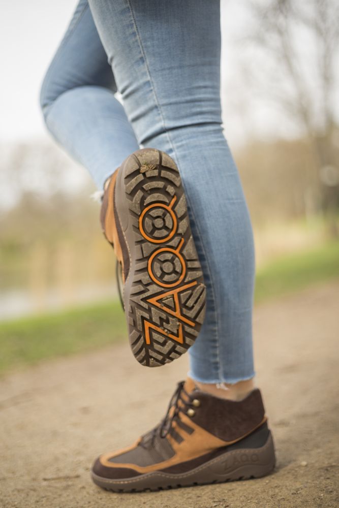 Eine Person in Blue Jeans hebt ihren Fuß und enthüllt die Sohle ihrer braunen ZAQQ SQOUT Waterproof Wanderschuhe, deren Veloursleder-Finish für einen Hauch von Stil sorgt. Im Hintergrund ist eine verschwommene Parkszene mit Bäumen und einem Pfad zu sehen.