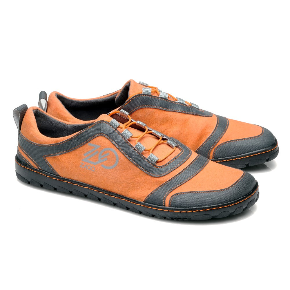 Wir präsentieren den SQILL Orange von ZAQQ: Diese sportlichen Sneaker im Barfußschuh-Stil zeichnen sich durch eine stilvolle Kombination aus Orange und Grau mit schwarzen Gummisohlen aus und präsentieren sich in einem eleganten, unauffälligen Design, das perfekt für moderne Minimalisten ist. Auf der Seite ist ein auffälliges stilisiertes Logo mit ZD zu sehen und ihre praktische schnürsenkellose Konstruktion sorgt sowohl für Komfort als auch für modische Attraktivität.